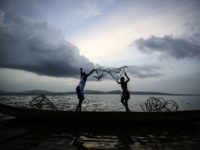 The silhouette of two people throwing a fishing net into a lake.