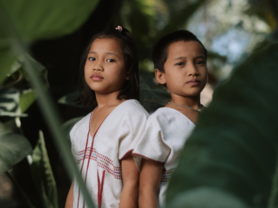 Two girls in white and red shirts stand amidst foliage, looking somber.