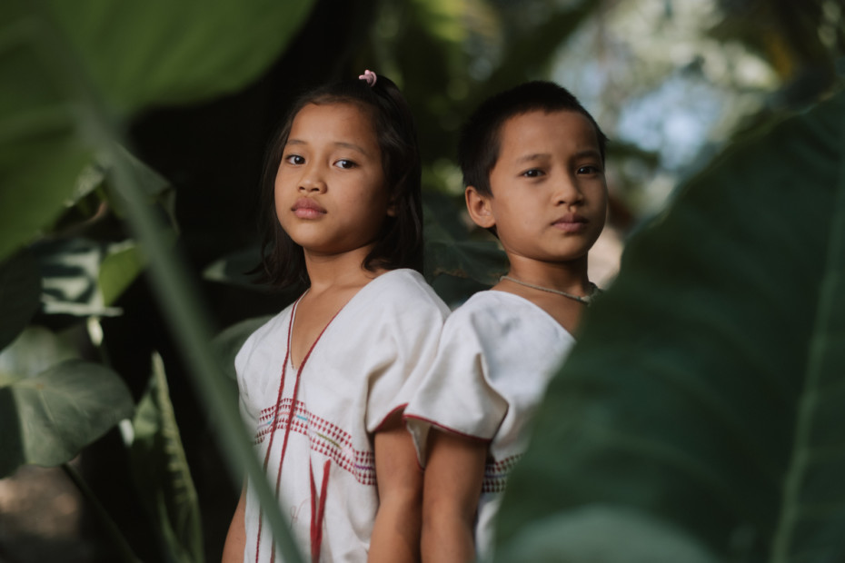 Two girls in white and red shirts stand amidst foliage, looking somber.