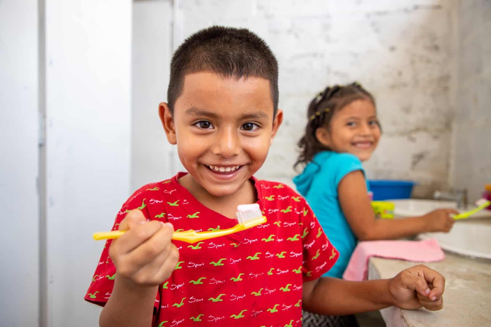 A boy in a red shirt holds up a toothbrush and smiles.