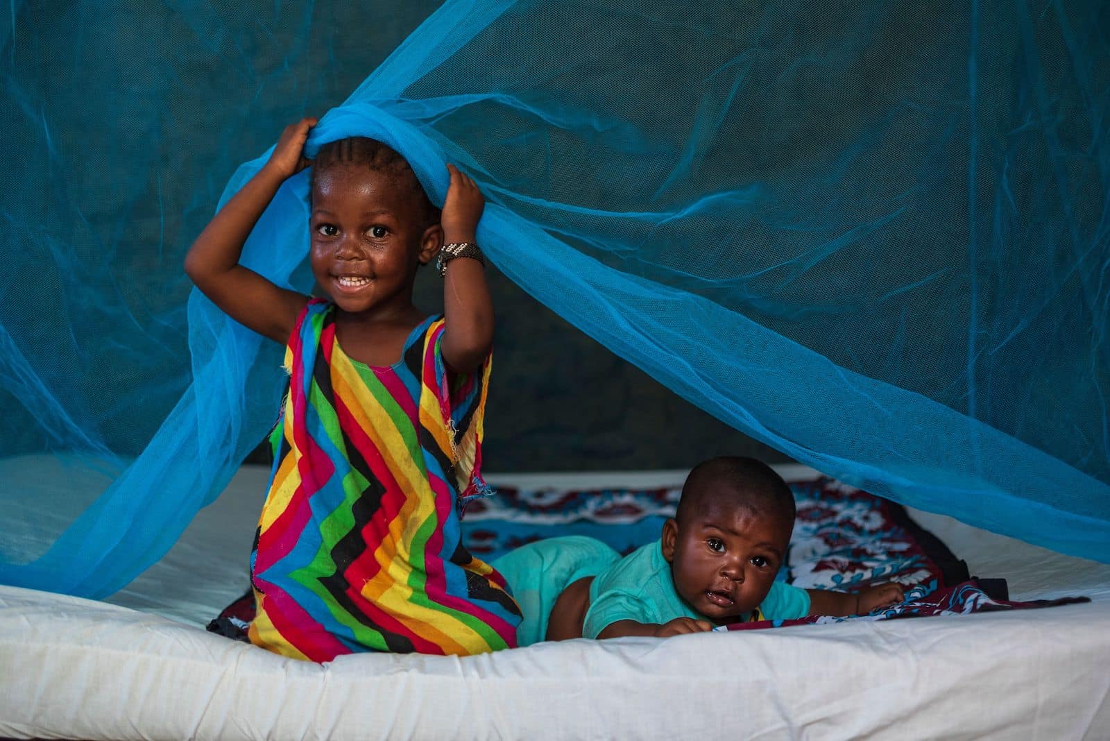 A girl and a baby sit on a bed with a malaria net on it. 