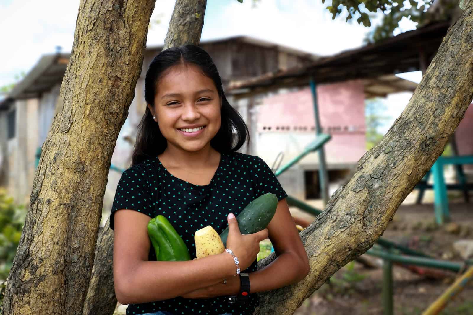 A girl leans against a tree, holding vegetables.