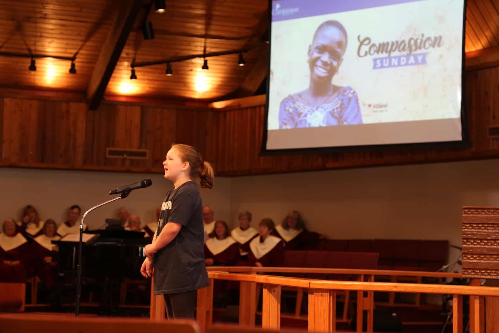 A girl stands on the stage of a church, speaking into a microphone.