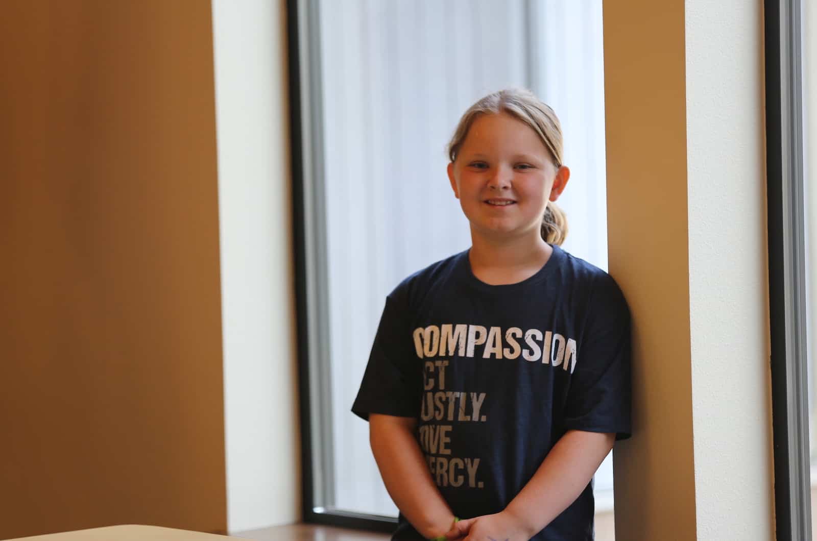 A girl in a T-shirt that says Compassion leans against a wall. 