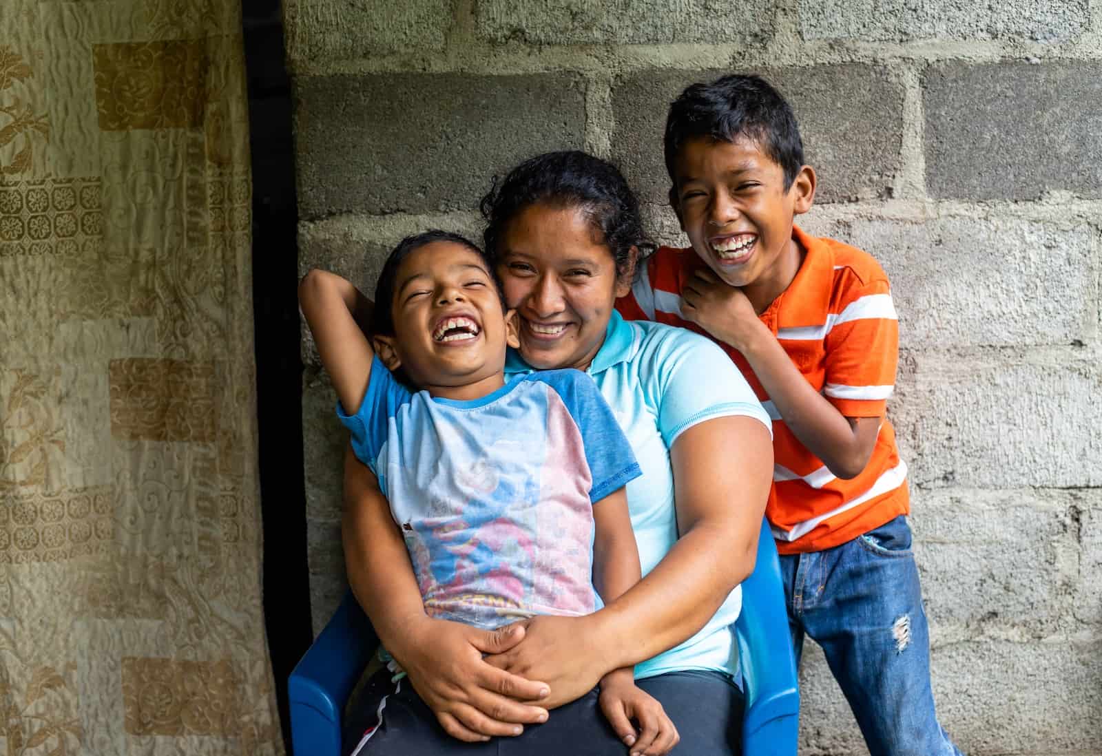 A woman holds a boy in her lap and a boy stands behind them, smiling.