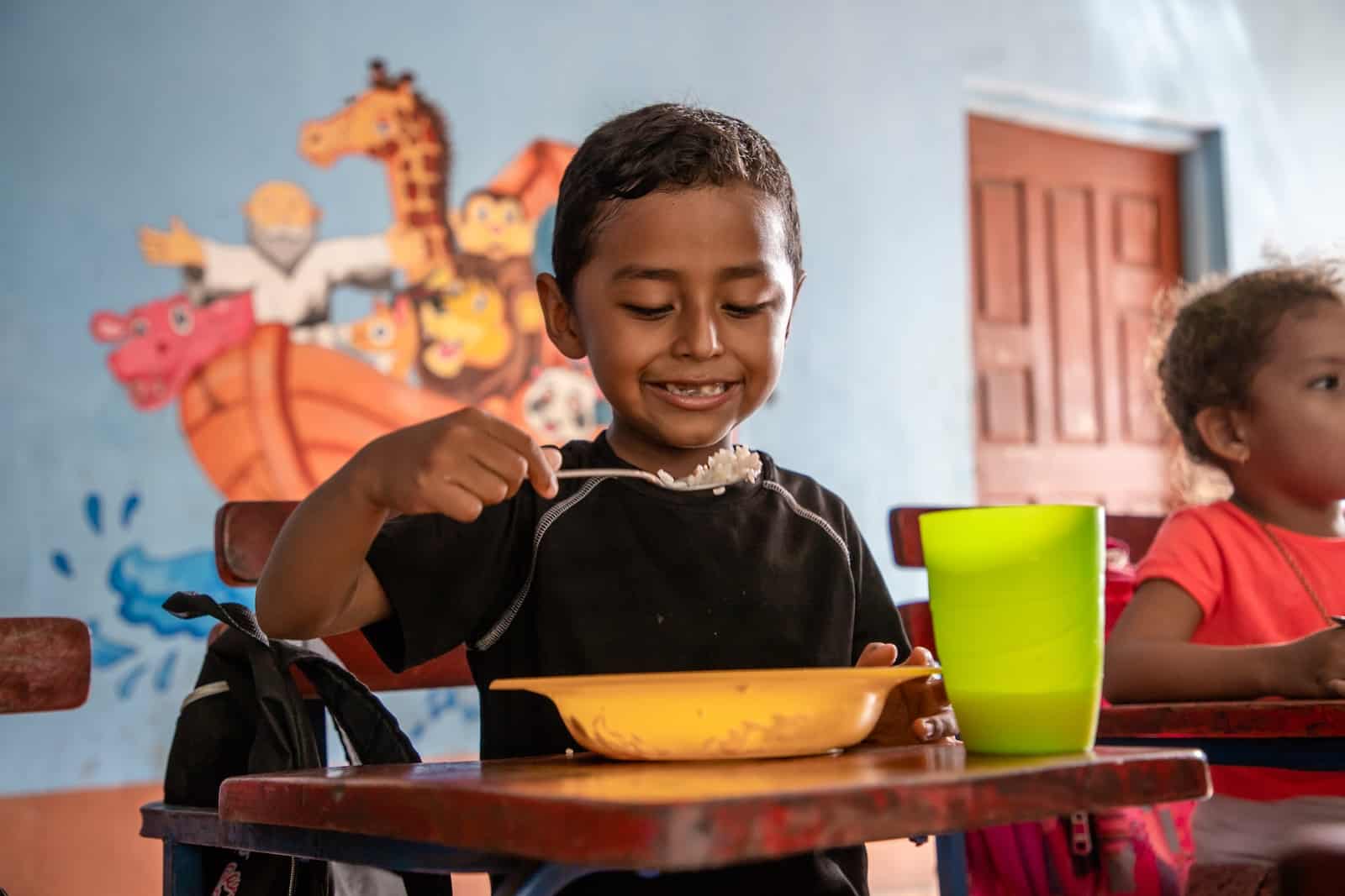 A boy sits at a desk, eating a bowl of food.