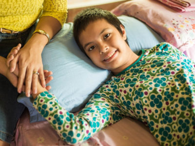 A girl lies in bed while someone holds her hand.