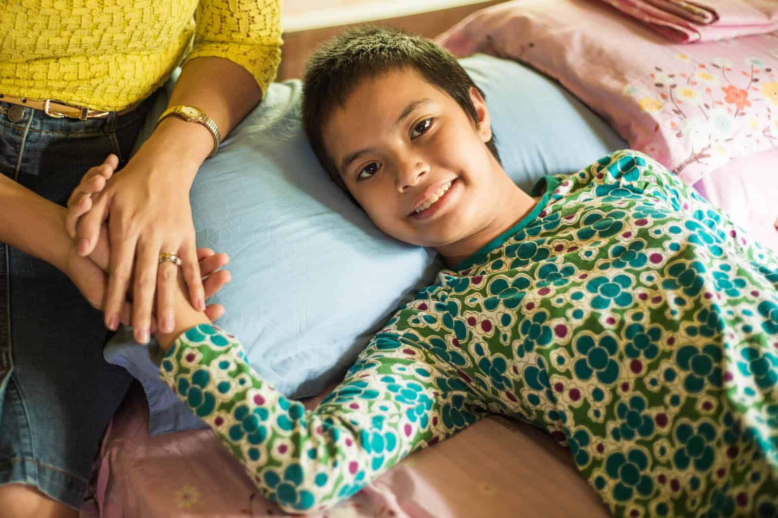 A girl lies in bed while someone holds her hand.