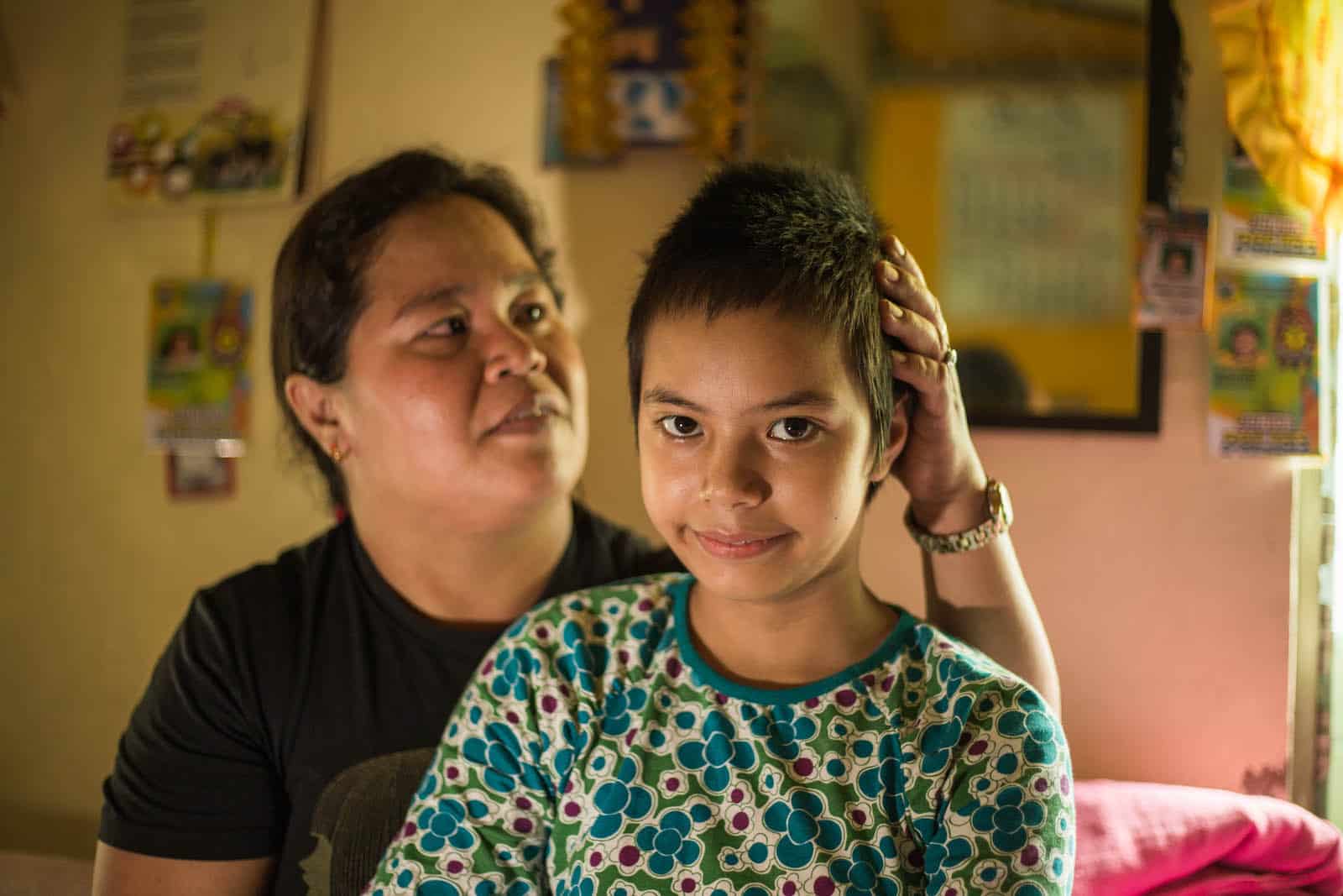 A girl with short hair sits in a room with her mother behind her.