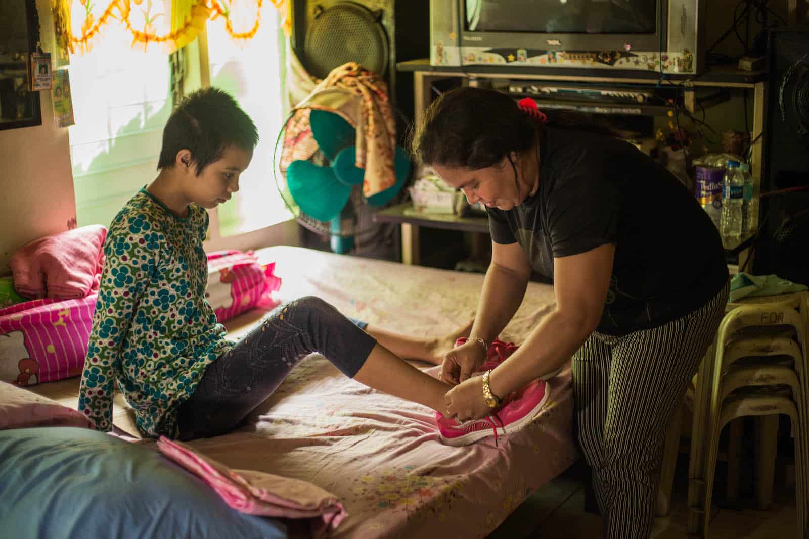 A woman ties a girl's shoes, sitting on a bed.