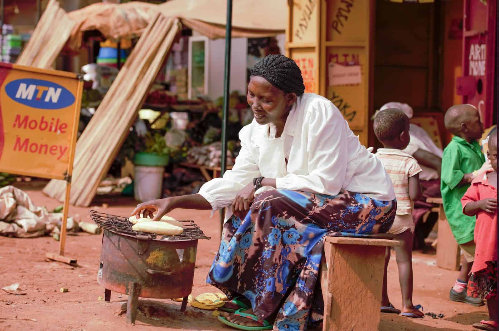 a woman sits on the side of a dirt road, cooking plantains on a simple woodstove.