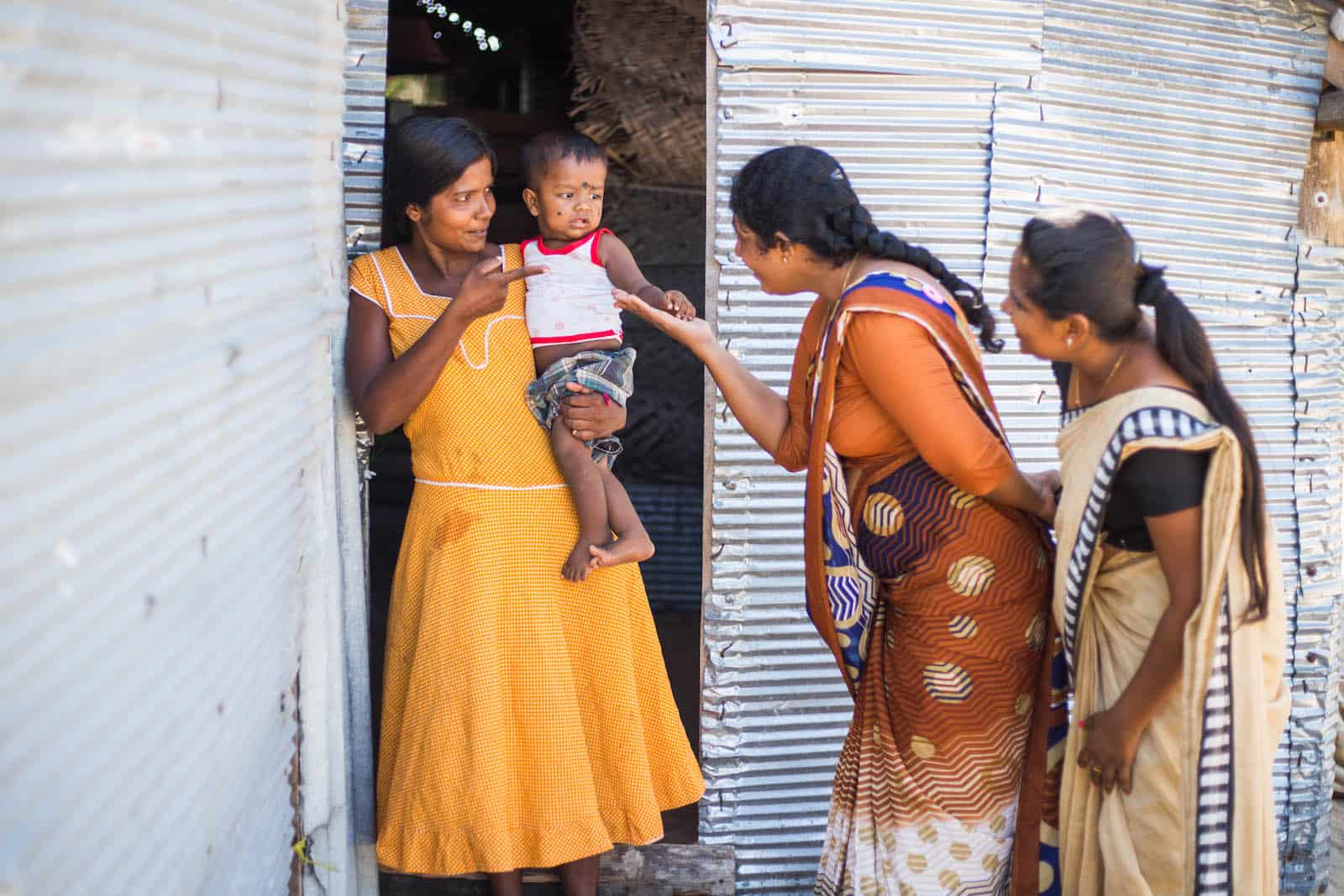 Visiting Sasiliya at Home Three women stand outside of a metal sheet home, one holding a baby.
