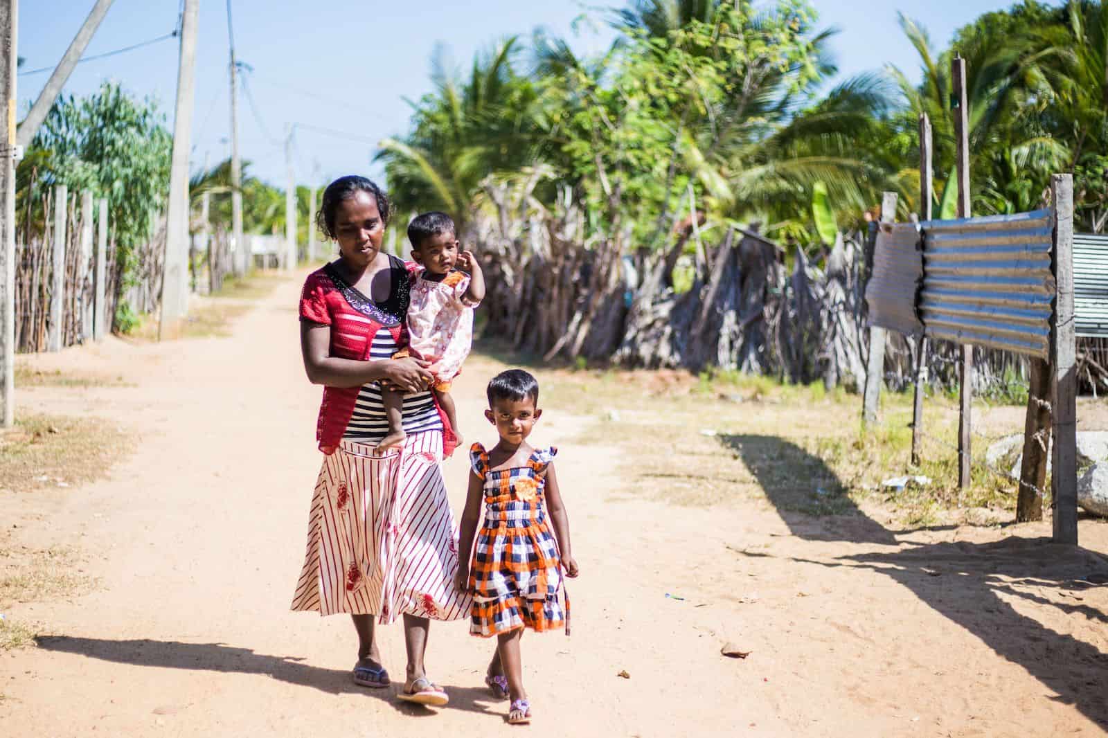 Udea and Her Children A woman holding a toddler and a girl walk down a dirt road.