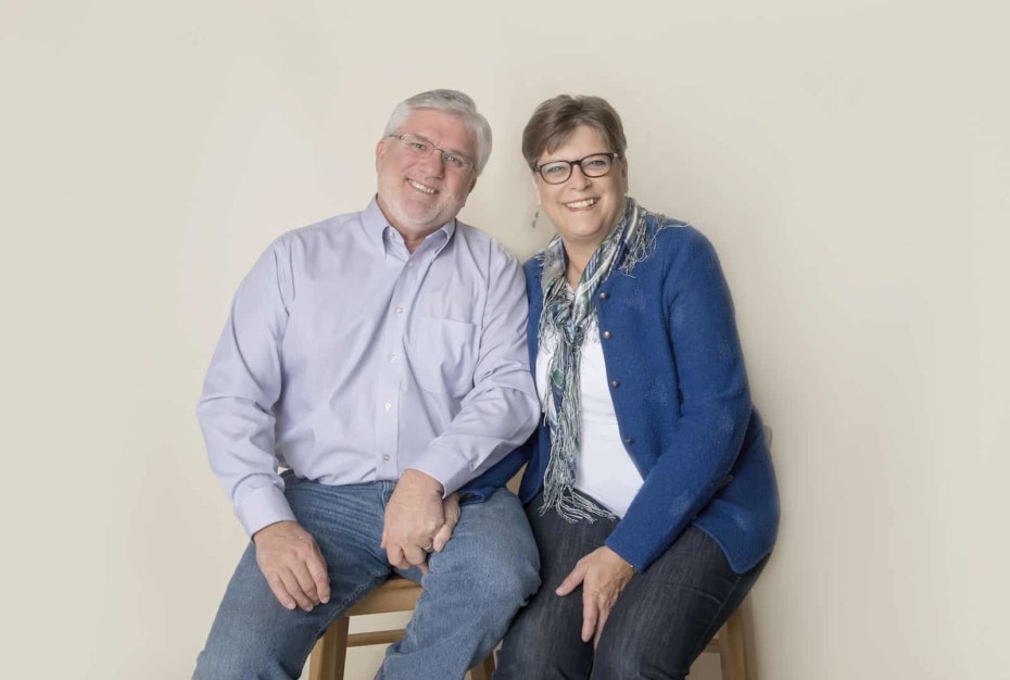 A man and woman sit on stools, holding hands.