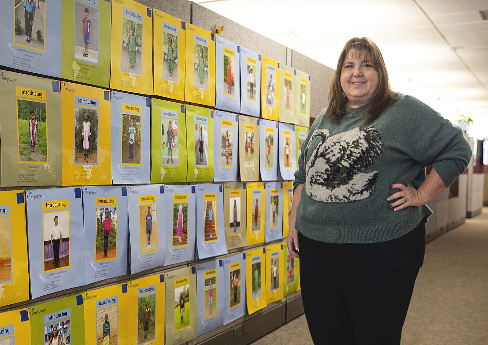 A woman in a green sweater stands in front of a wall covered in pictures of children.