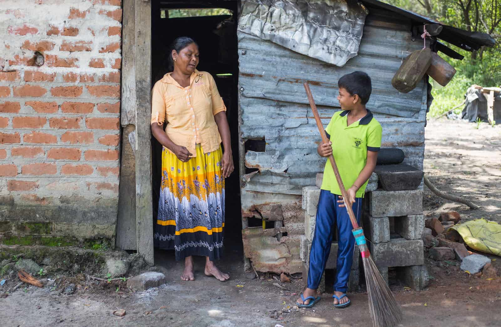 A boy sweeps in front of a metal sheet and brick home while a woman watches.