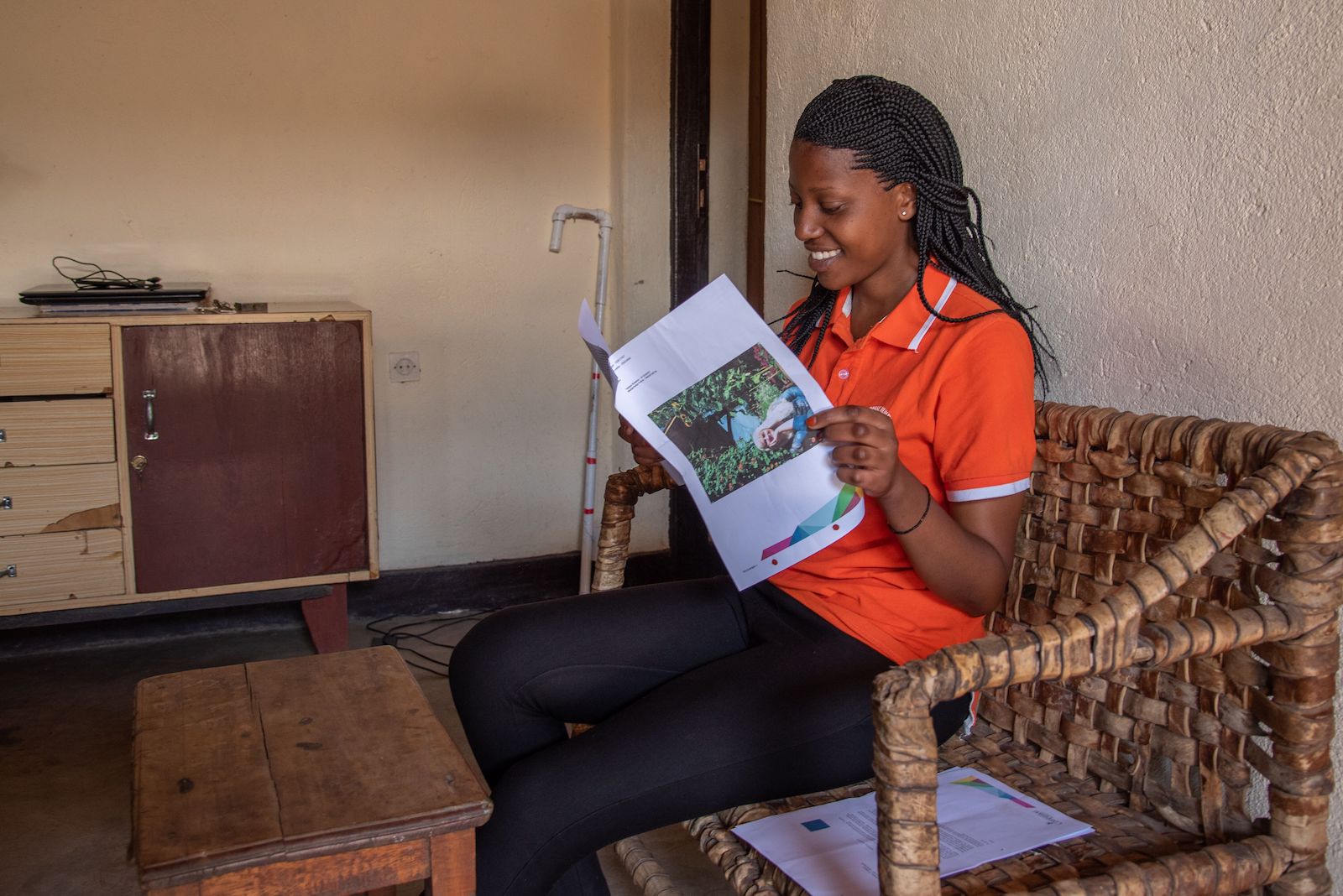 A young woman in an orange shirt sits, reading a letter. 