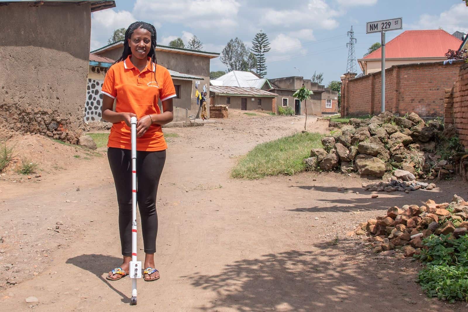 A girl in an orange shirt stands on a dirt road, holding a blind stick.