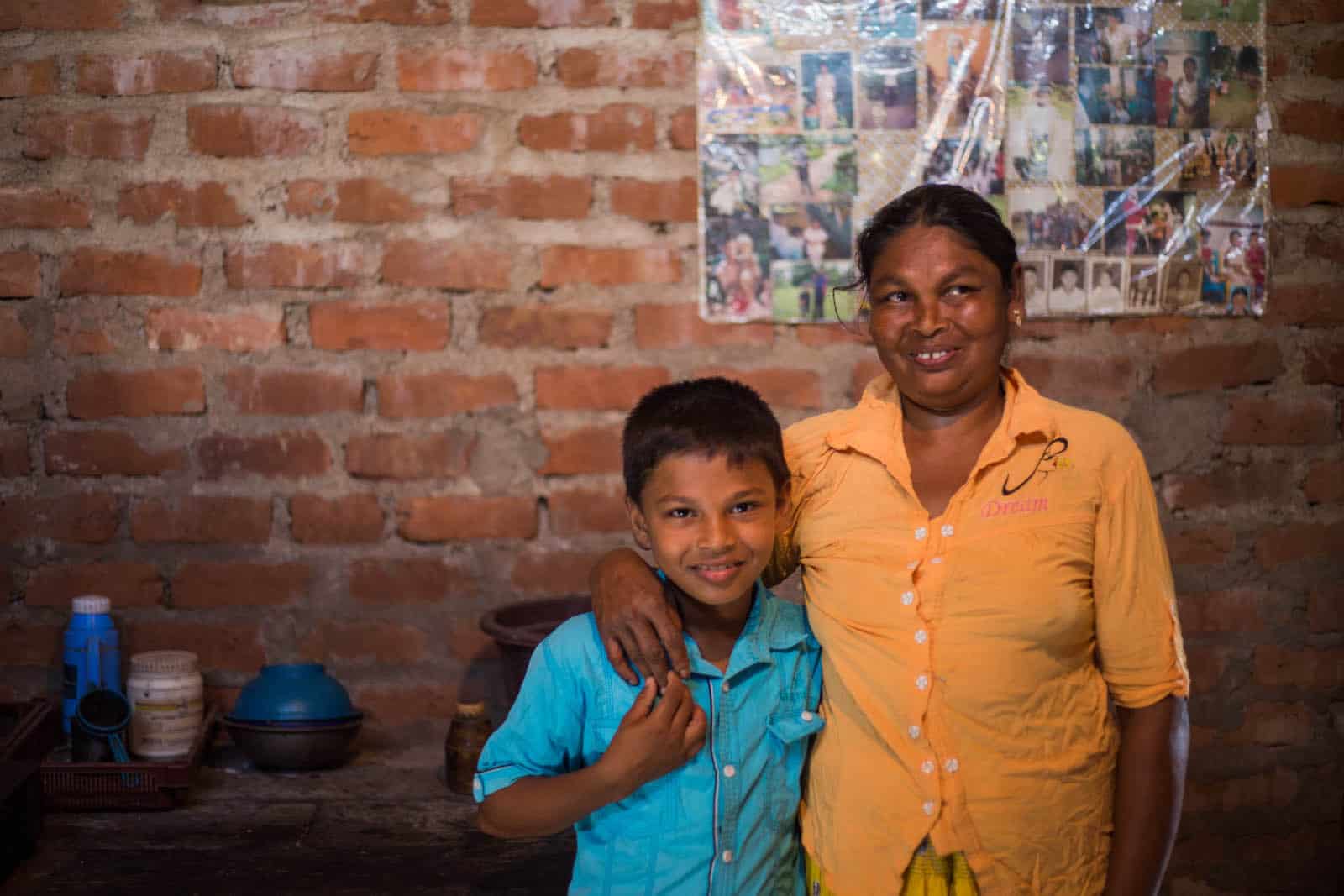 A boy stands next to a woman, smiling, standing in front of a brick wall. 