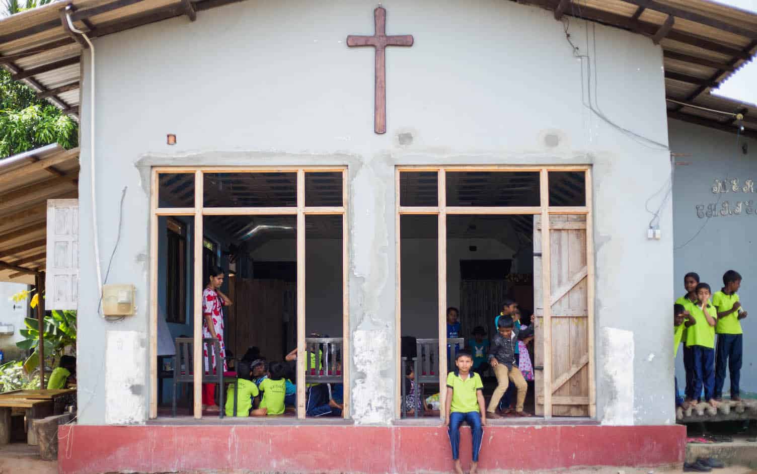 A boy sits in front of a church with large windows and a cross.