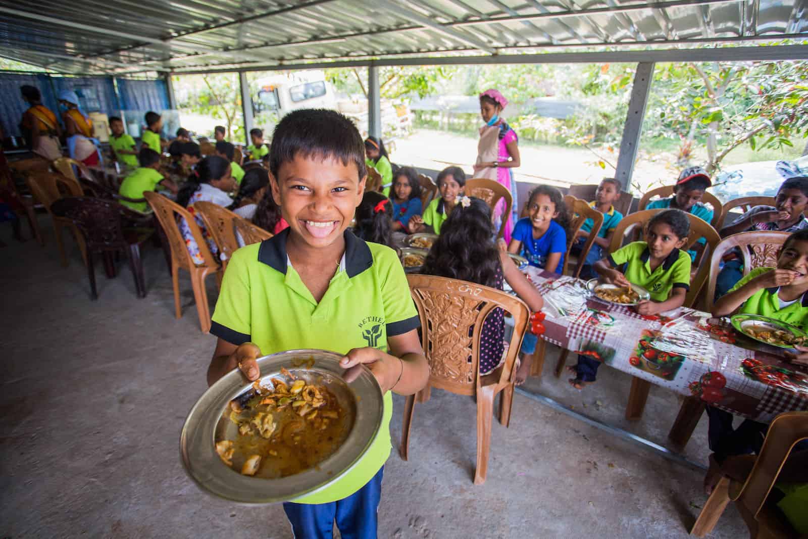 A boy holds out a plate of food, standing in front of a table of children eating.