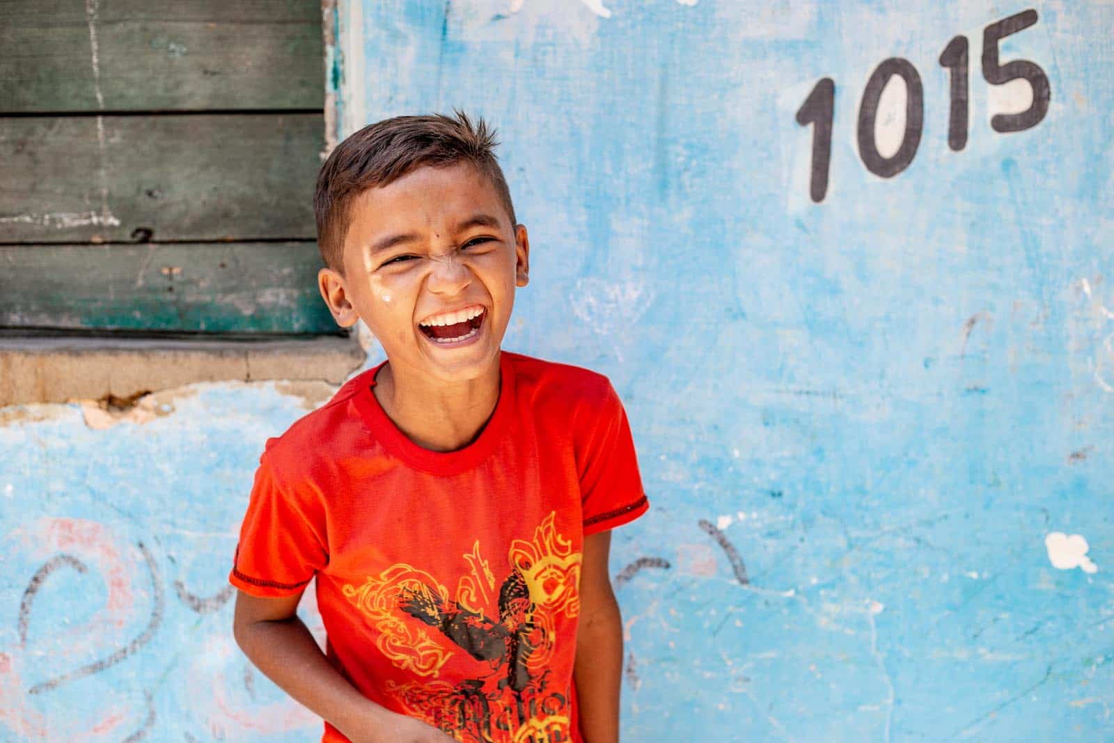 A boy in a red shirt laughs at the camera.