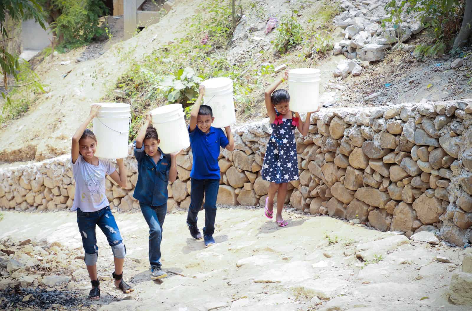 Four children walk up a hill carrying water buckets
