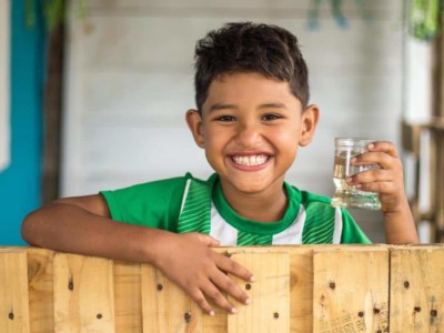 A boy stands at a fence holding a glass of water.