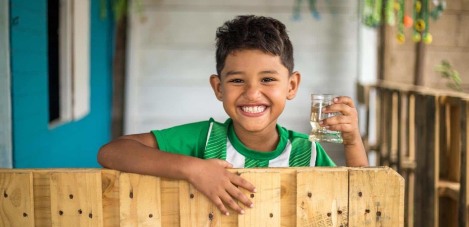 A boy stands at a fence holding a glass of water.