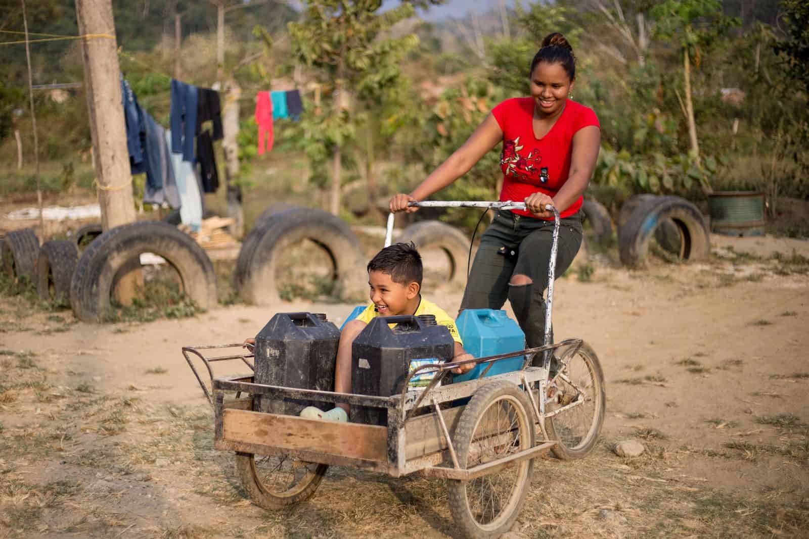 A boy rides in a tricycle cart holding water jugs, as a woman pedals.