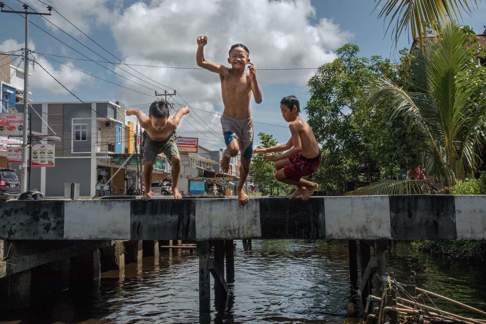 Kids Around the World: Three boys jump into a ditch next to a city block.