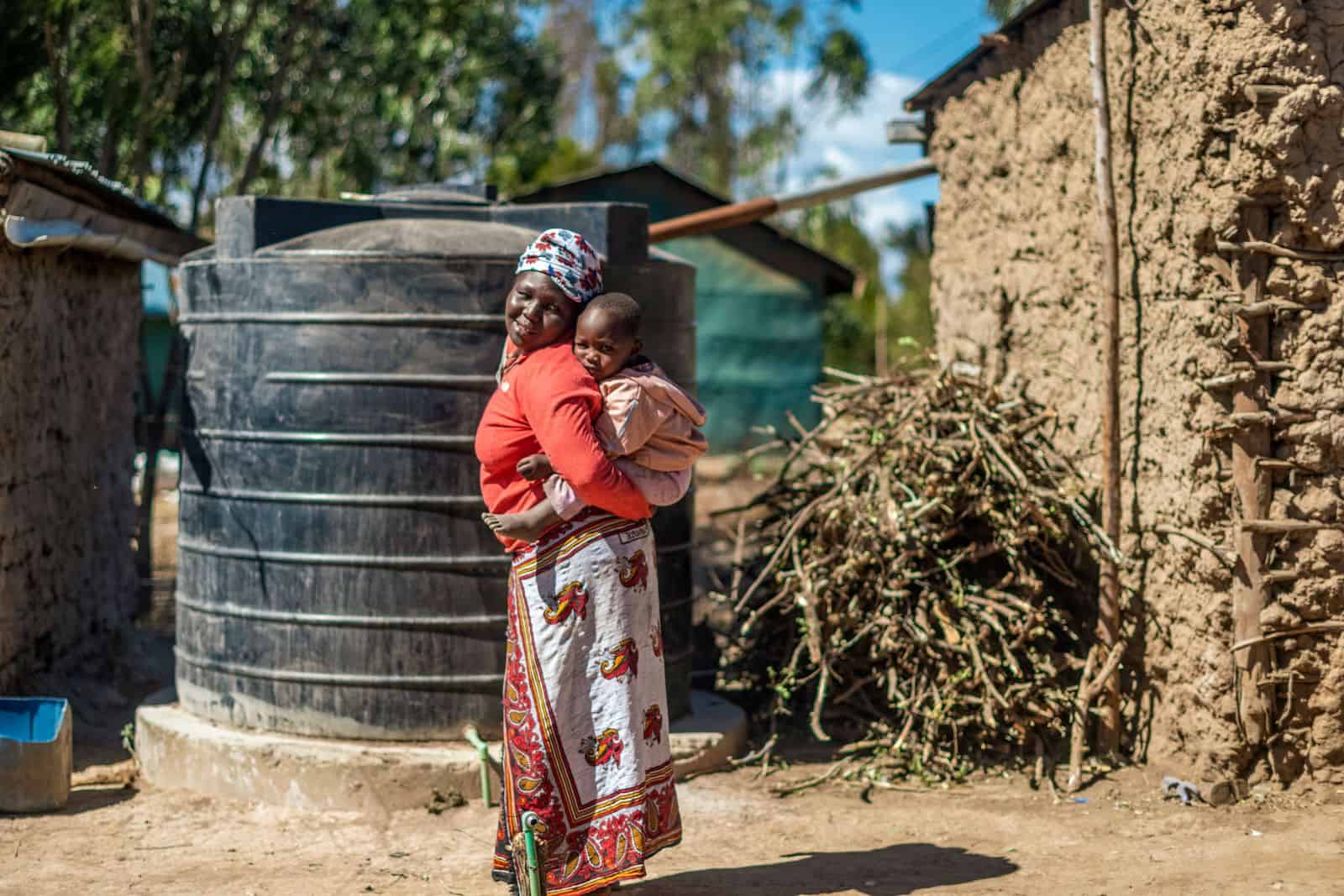 A woman with a baby on her back stands in front of a mud house with a water tank.