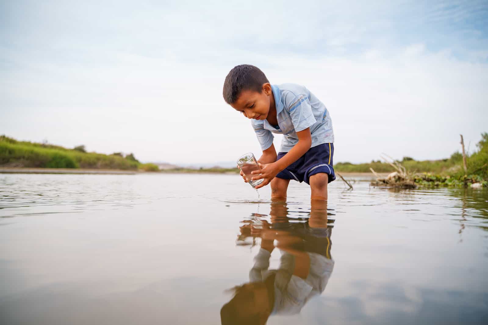 A boy leans over a river with a cup.