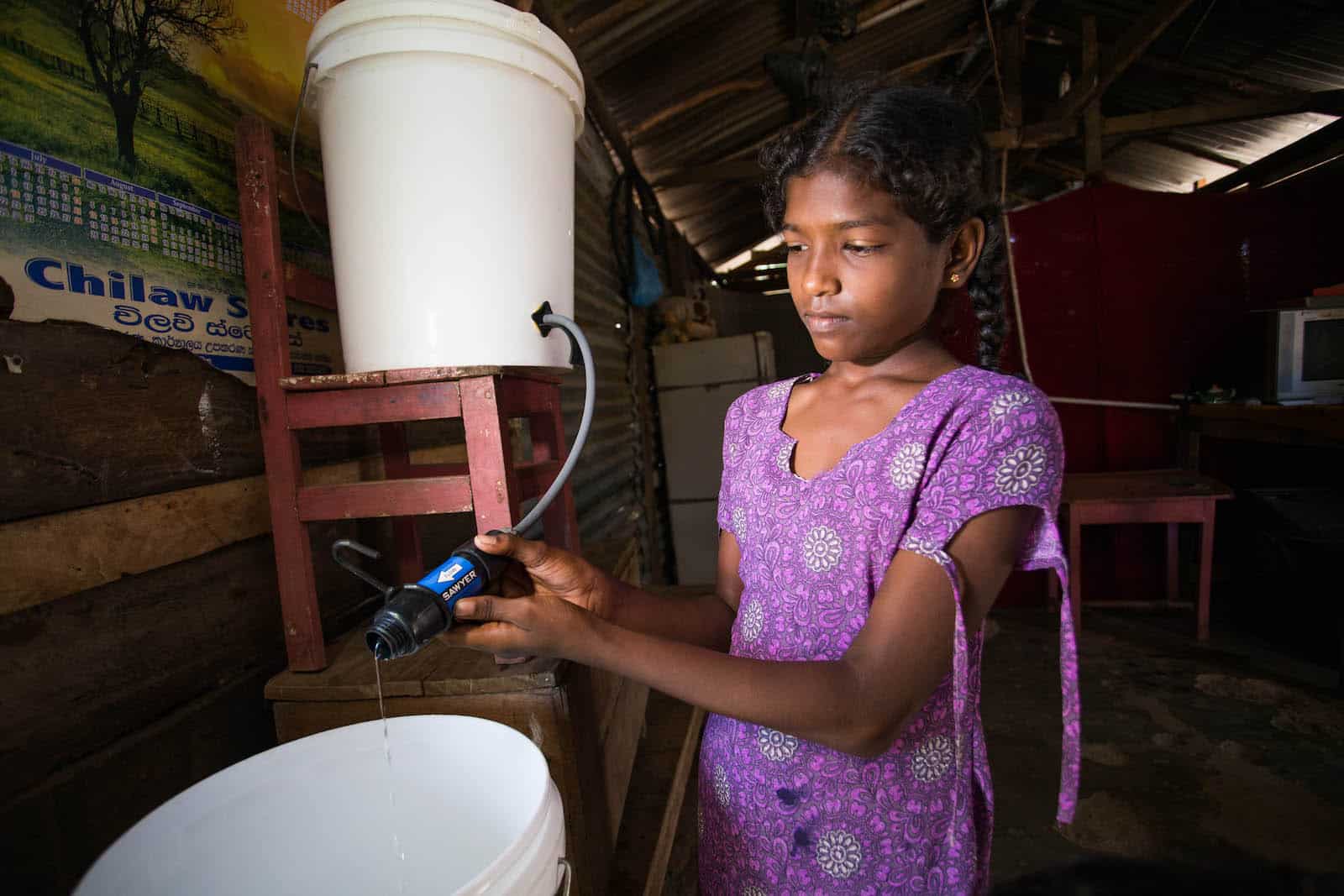 A girl in a purple shirt stands inside holding a water filter connecting one bucket to another.