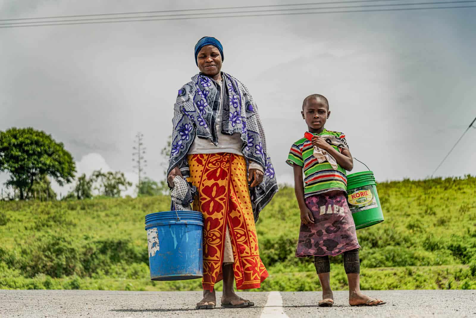A woman and a girl stand on a street, holding buckets