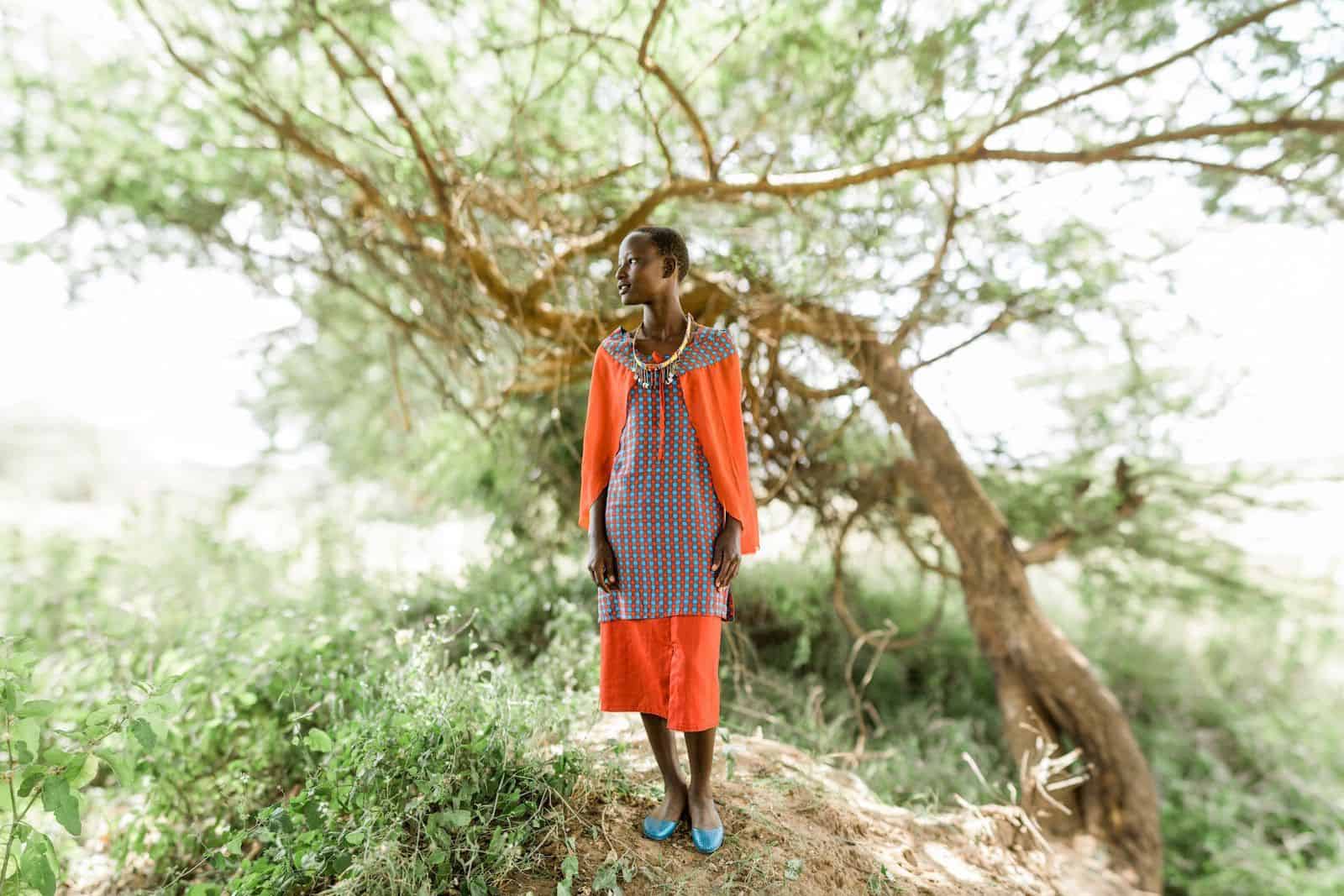 A girl in an orange dress stands in front of a leaning tree. 