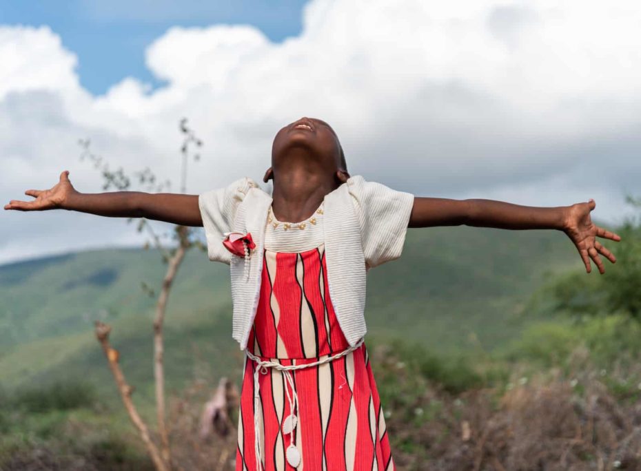 A girl in a red dress stands in the hills, head back and arms out to the side.