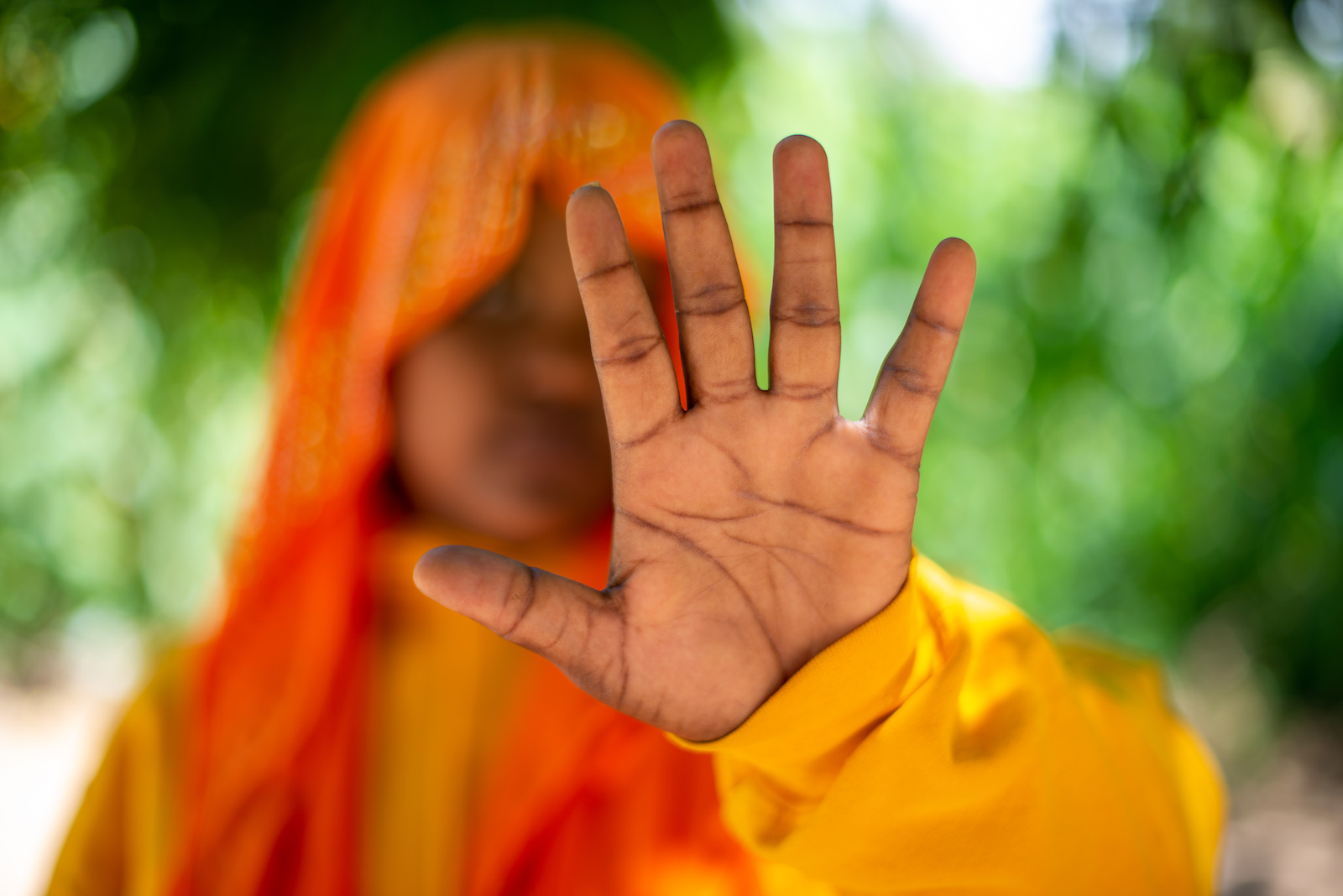 A girl in orange wearing a head covering holds her hand in front of a camera to block her face.