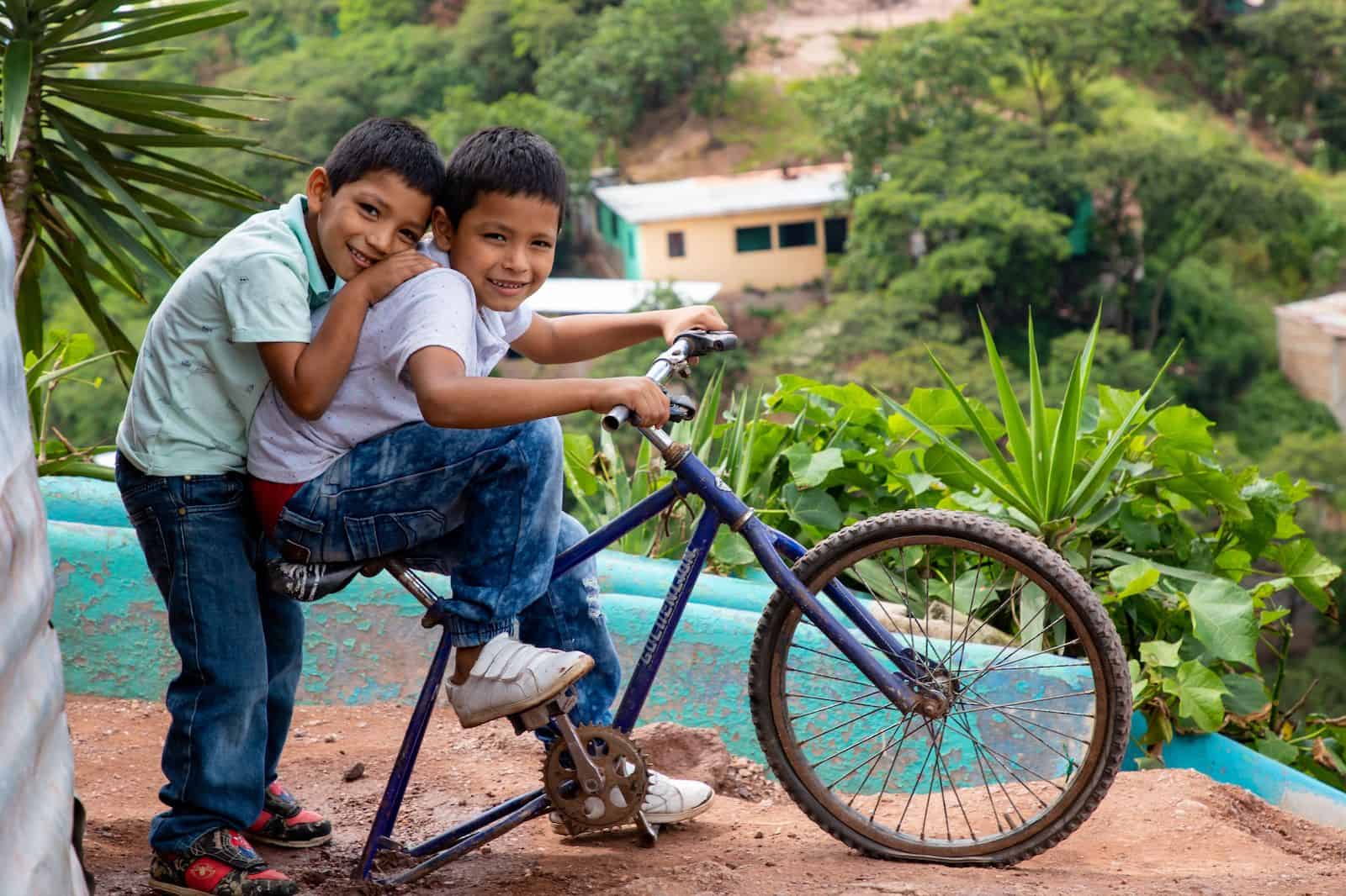 A boy sits on a bike with a boy behind him, hugging him.