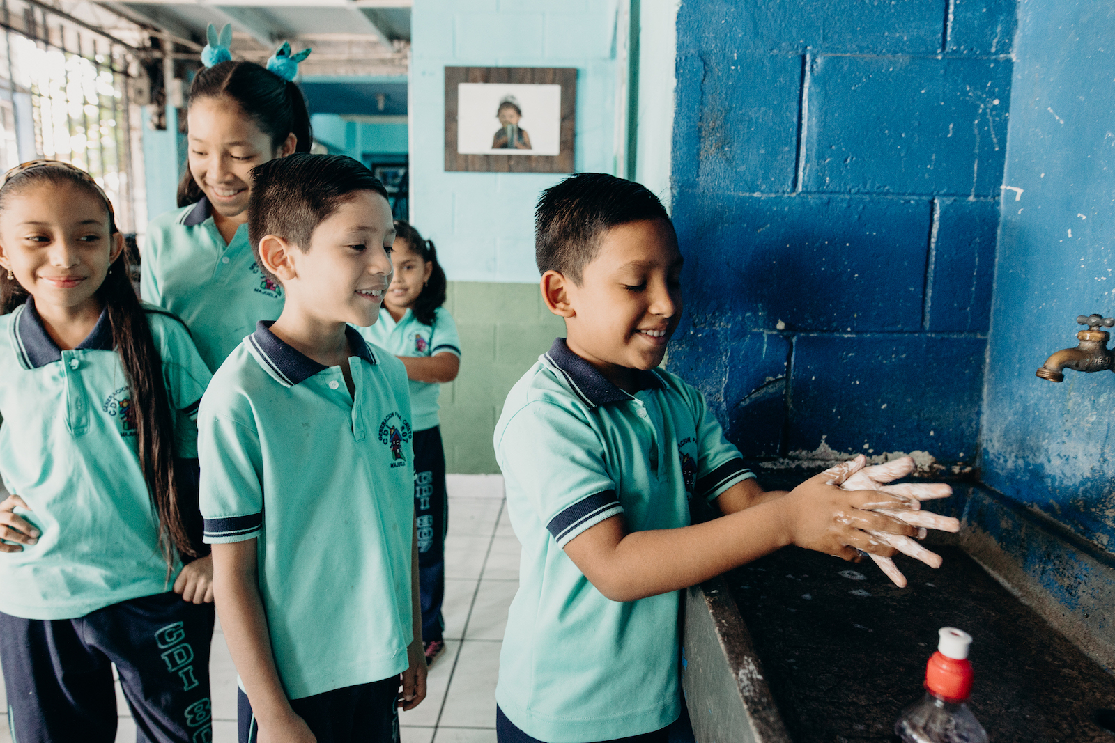 A boy washes his hands while children wait behind him.