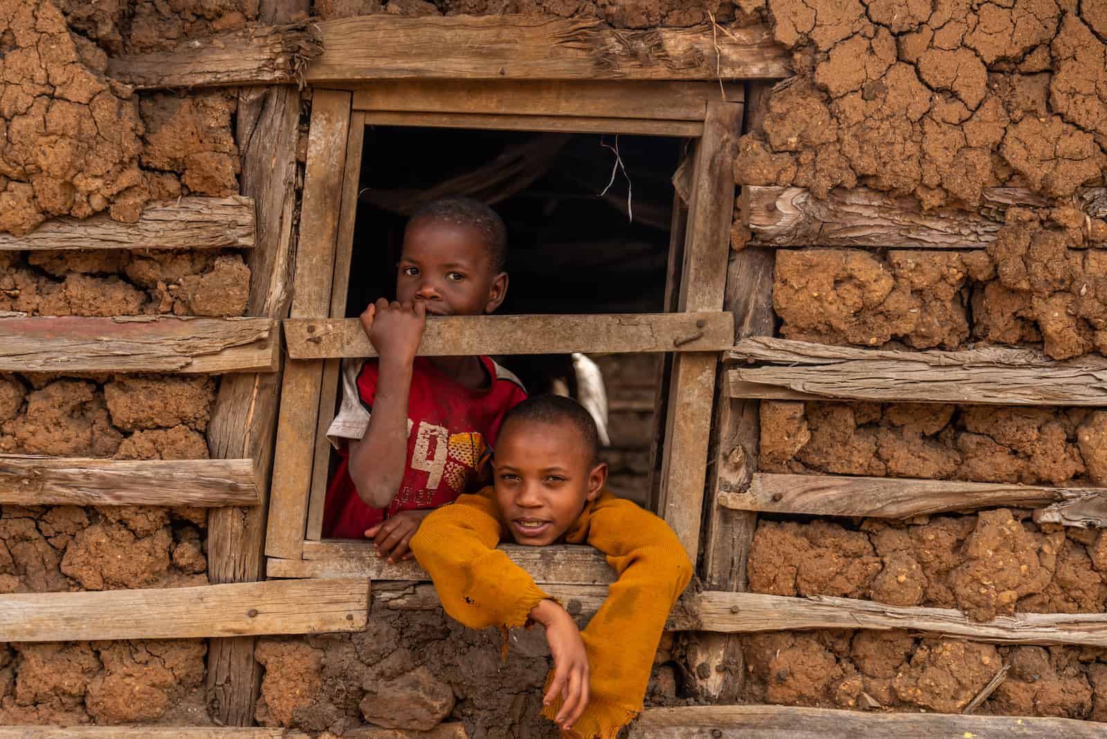Two boys look out the window of a mud and wood home.