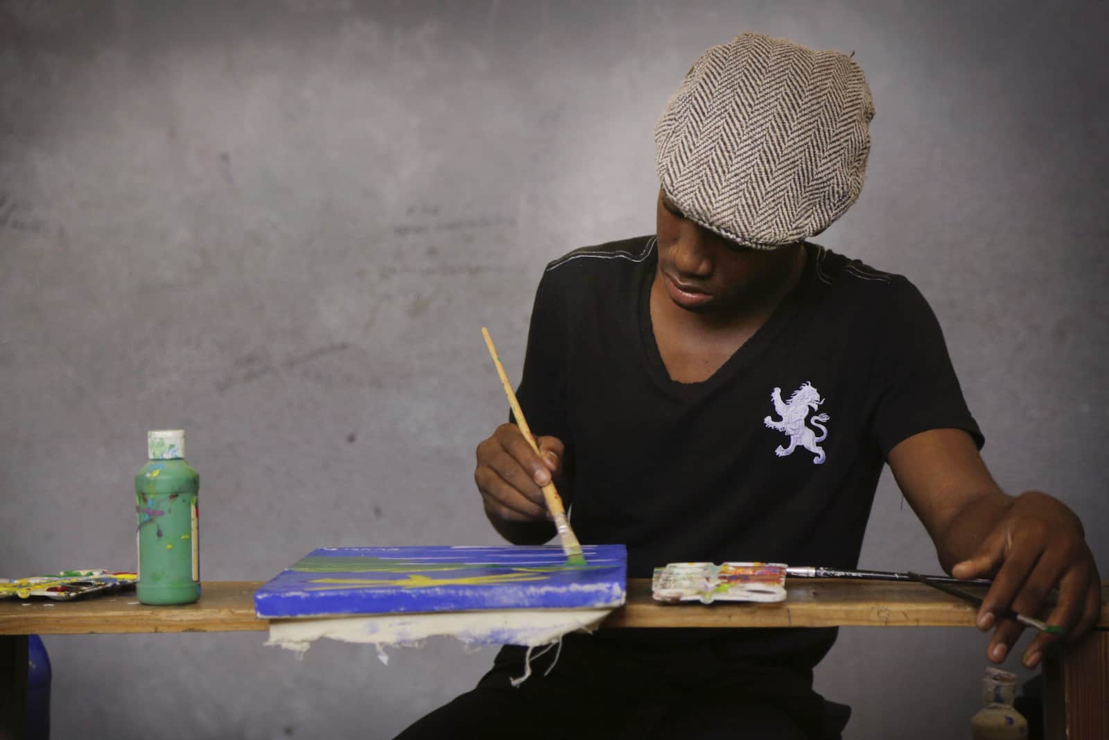 A man in a grey beret sits at a table, painting.