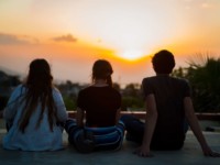 Three people sit cross-legged in front of a sunrise.