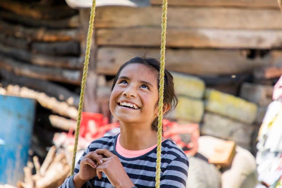 A girl sits on a swing, looking up and smiling.