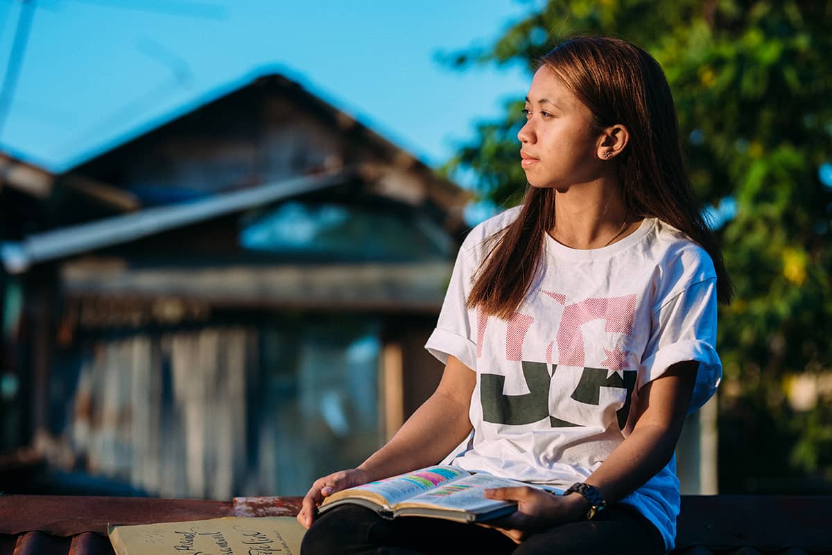 A girl, in a white shirt and black pants, is sitting outside with a Bible open in front of her on her lap. There is a building and a tree in the background.