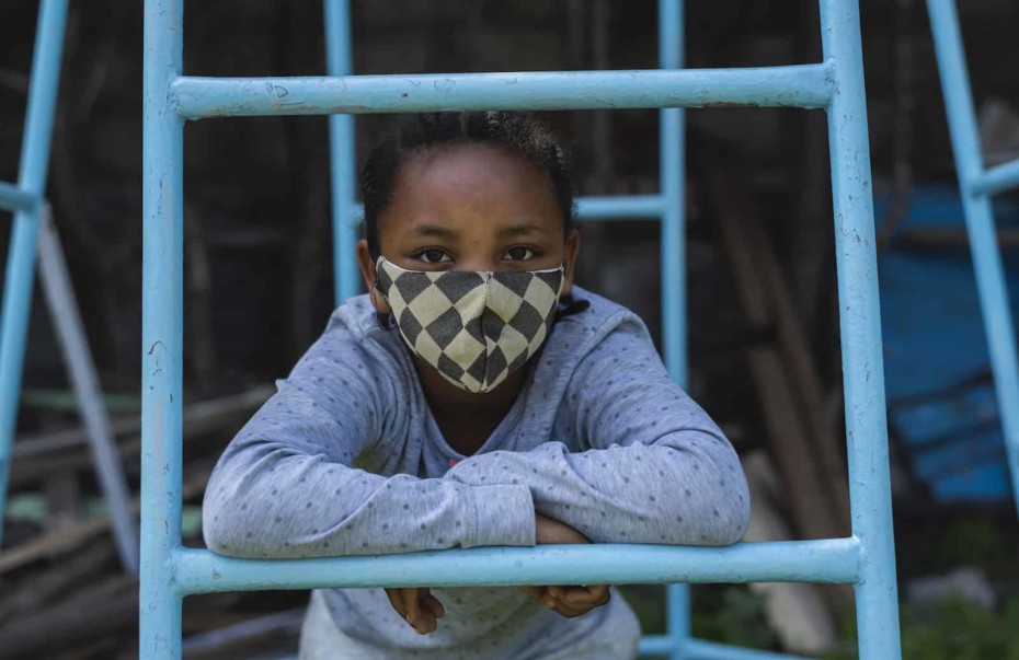 A girl with a checked face mask leans on a jungle gym.