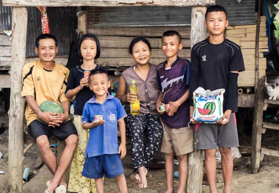 A family of six sits outside a wood home holding food.