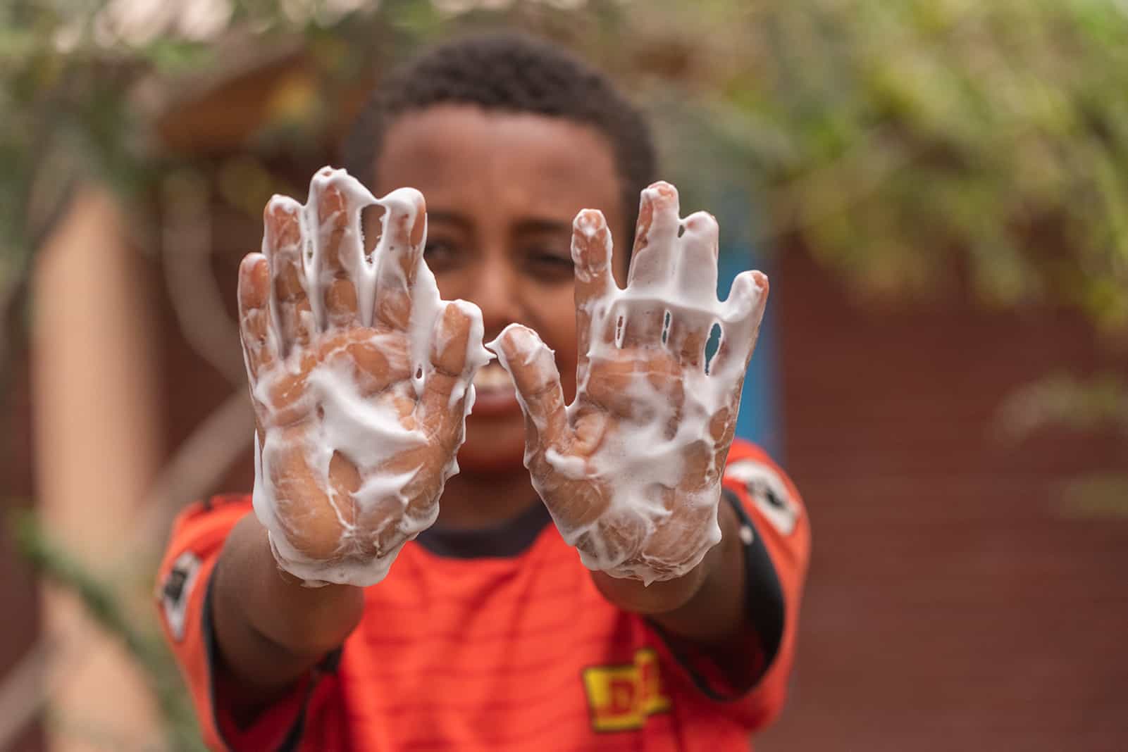 A boy wearing a red shirt holding his lathered hands up to the camera