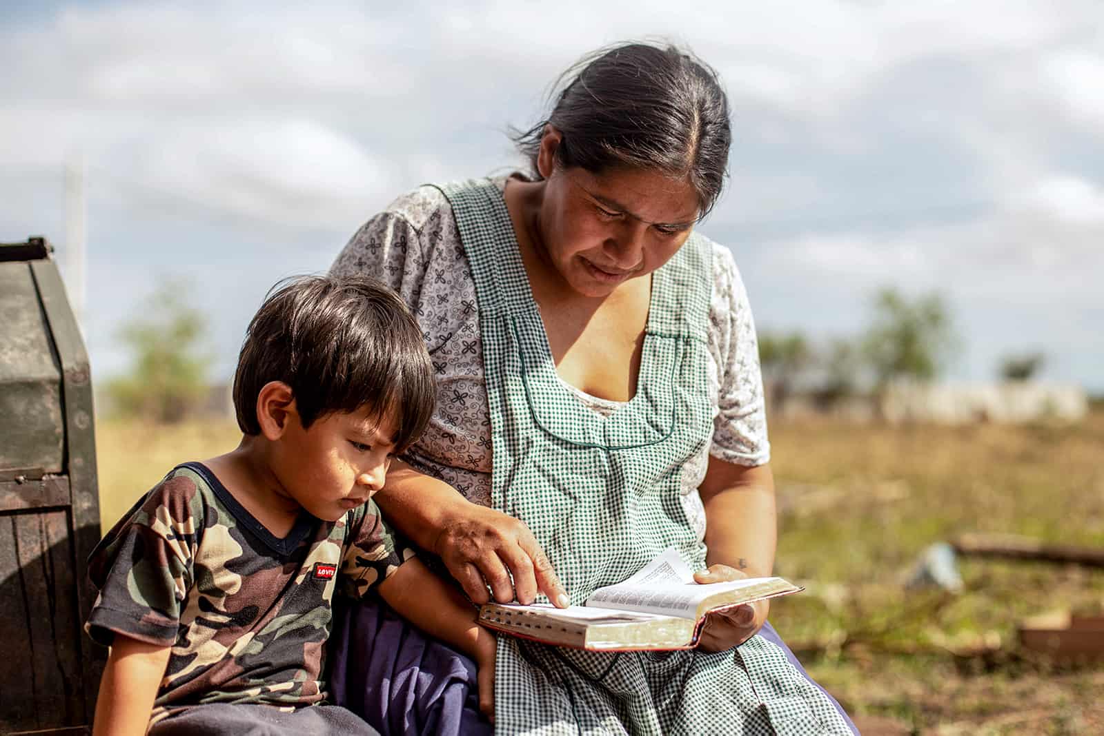 Andy is sitting next to his mother while she reads. Andy is wearing a camoflauge shirt and his mother is wearing a blue and white patterned shirt.