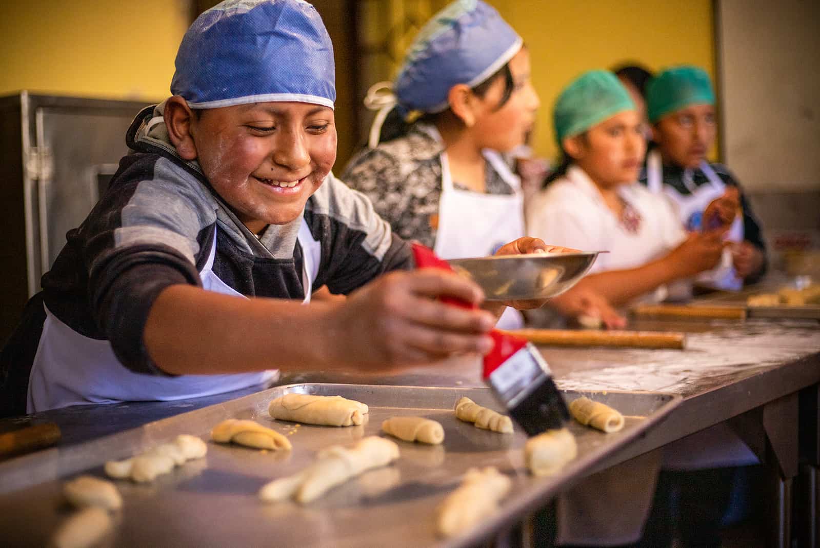 Juan, in a white apron and blue head covering, is using a paint brush to brush egg onto rolls of bread dough that are on a pan on the table in front of him.